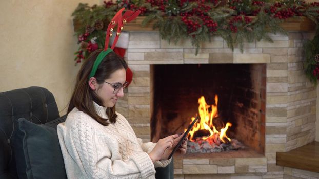 A young pregnant woman with glasses sits in a chair by the fireplace with horns on her head and writes on the tablet. 4k
