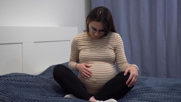 Young pregnant woman with glasses stroking her belly. The girl in the bedroom on the bed. The camera zooms in. 4k
