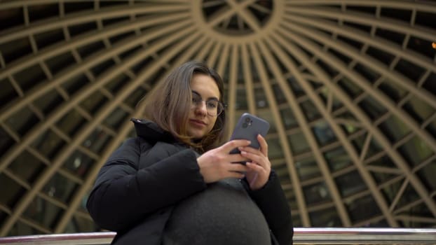 A young pregnant woman in glasses and a jacket stands with a phone in her hands. A girl in a shopping center uses a smartphone against the backdrop of a panoramic dome. 4k