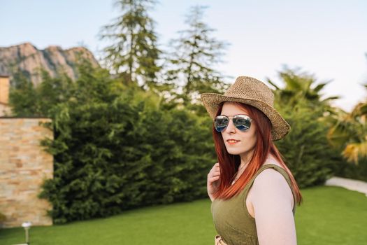 Girl with straw hat and sunglasses dressed in summer clothes. smiling woman on summer holiday in a hotel. outside with natural light, background with garden vegetation, clear sky and mountains.