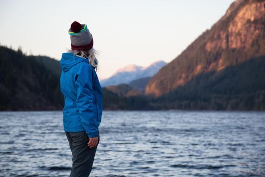 Young girl wearing a tuque and a jacket is watching a beautiful mountain scenery during sunset. Taken at Buntzen Lake, Vancouver, British Columbia, Canada.