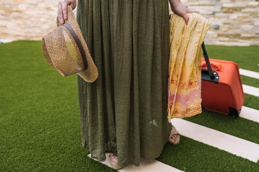 cropped shot of a young woman, entering the hotel reception with her suitcase on wheels. woman on a pleasure trip dressed in summer clothes. Outside, dim sunlight, grass floor with white tiles.