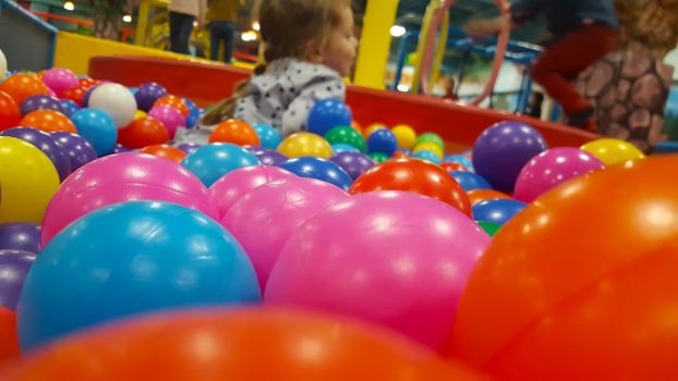 Happy cute girl having fun in ball pit in kids amusement park and indoor play center. Close up portrait child playing with colorful balls in playground ball pool. Activity toys for little kid.