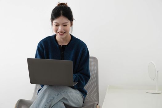 portrait of a happy casual asian woman holding laptop computer while sitting on a chair over white background.