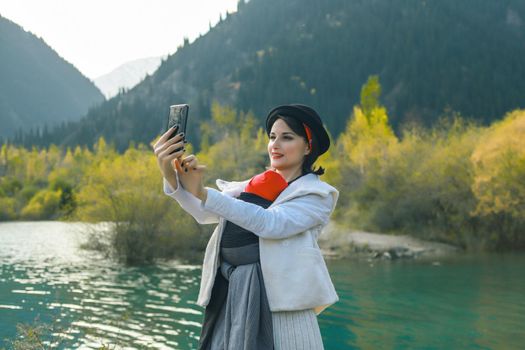 Beautiful young mother with her infant baby in sling make selfie. Mother is carrying her child and travel in autumn mountain lake. Active mother concept