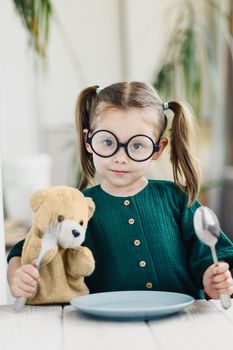 Child waiting breakfast with bear toy. Little cute girl at white dining table in kitchen. Healthy nutrition for young kids.