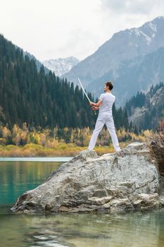 A man in white clothes with a sword stands on a large stone among the highlands