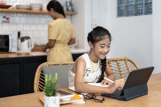 Portrait of little girl using laptop during online class with tutor or teacher while sitting at desk in cozy kitchen interior with mother