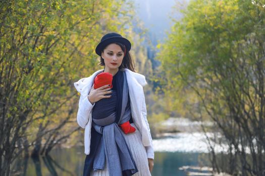 Beautiful young mother with her infant baby in sling outdoor. Mother is carrying her child and travel in autumn mountain lake. Babywearing concept