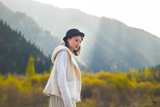 Beautiful woman in a white coat and black hat posing on a background of autumn mountains