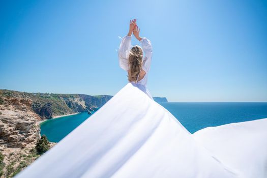 Blonde with long hair on a sunny seashore in a white flowing dress, rear view, silk fabric waving in the wind. Against the backdrop of the blue sky and mountains on the seashore