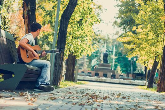 Young man sitting on the bench in the park playing on acoustic guitar with capo. Young attractive man enjoys live music in last sunny days autumn holiday. Retro lens used.