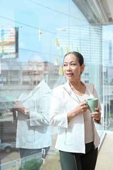 Thoughtful senior female business leader holding coffee cup and standing near large window at office with city buildings in background.