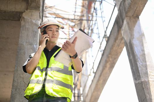 Young engineer man in vest and safety helmet talking on mobile phone under new building.