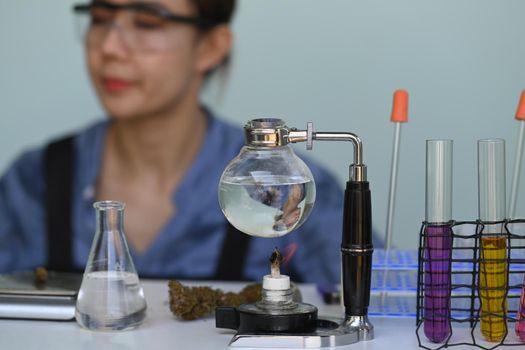 Glass bowl with cbd oil extracted from a marijuana plant on white table and female scientist conducting experiment in background. Herbal alternative concept.