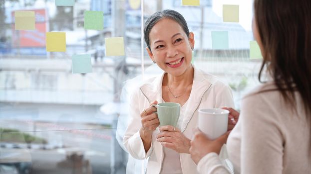 Positive middle aged female team leader standing near large window and having conversation with young employee during coffee break.