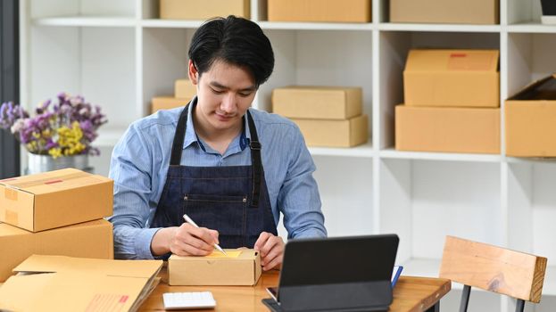 Young man online seller writing address on cardboard box while prepare parcel box of product for deliver. Concept of online selling, e-commerce.