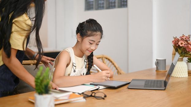Smiling asian girl doing assignments, studying online with her mother in home kitchen.