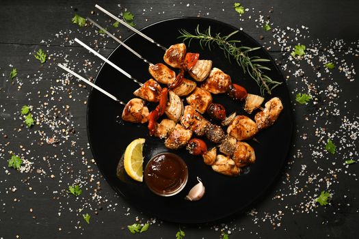 Plate with grilled chicken meat skewers and barbecue sauce on black rustic wooden table.