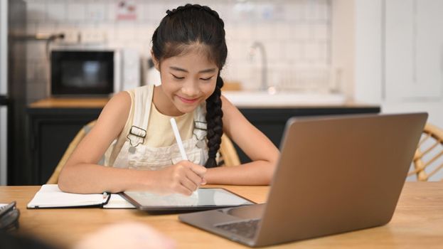 Cute Asian girl distance learning online, doing homework while sitting at kitchen table.