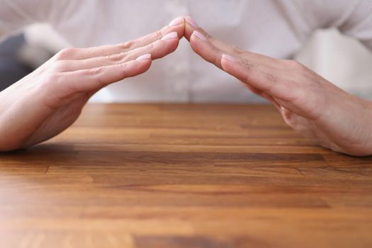 Close-up of female sitting at wooden table and forming hands house roof symbol with hands. Home safety and property security. Real estate insurance concept