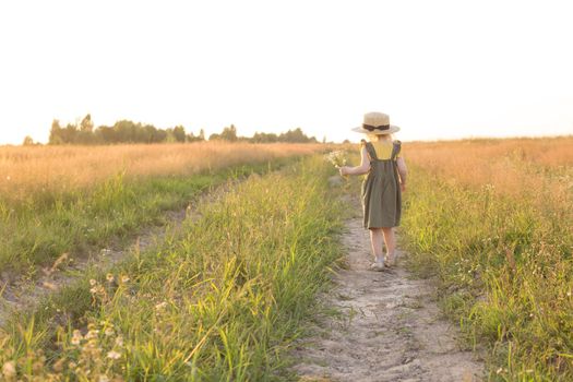 A little blonde girl in a straw hat walks in a field with a bouquet of daisies. The concept of walking in nature, freedom and an eco-friendly lifestyle.