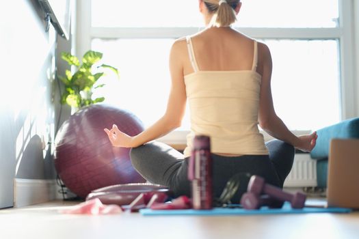 A woman sits in front of a window in a yoga pose, view from the back. Calm exercise, pandemic fatigue, self-isolation