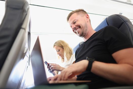 Close-up of happy man working on laptop, calm and joyful on airplane trip, woman use smartphone for entertainment. Remote job, freelance, technology, high in sky concept