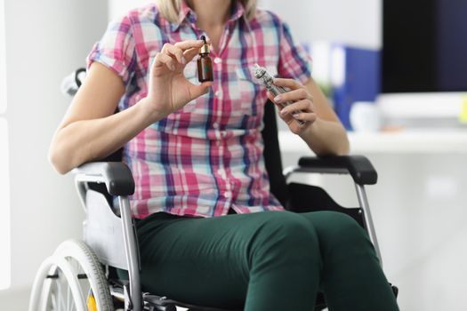 Close-up of person in disabled carriage in clinic. Woman holding glass bottle with cannabinoid oil and cannabis sign. Spinal fracture rehabilitation. Medical marijuana for treatment