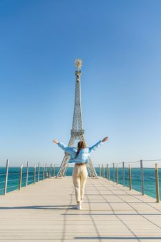 Large model of the Eiffel Tower on the beach. A woman walks along the pier towards the tower, wearing a blue jacket and white jeans