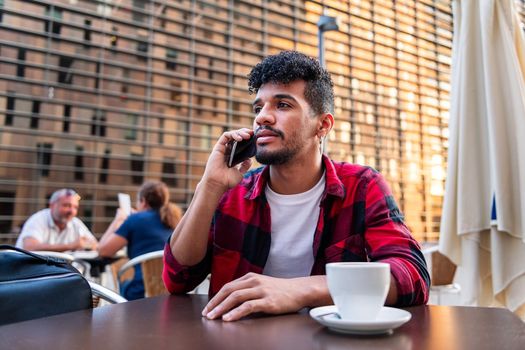 young latin man talking on the phone while sitting with a coffee on the terrace of a coffee shop, concept of technology and urban lifestyle