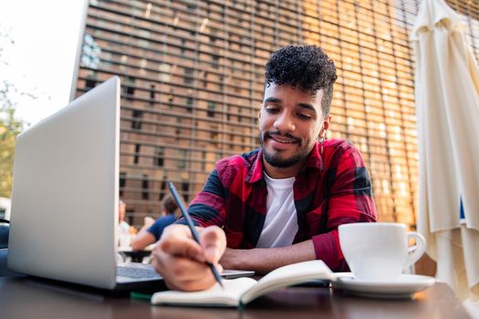 young latin student works with his computer and takes notes on the terrace of a coffee shop in the city, concept of technology and urban lifestyle