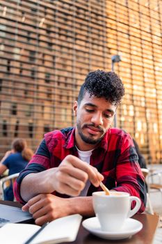 vertical photo of a young latin man stirring his coffee with a spoon while working on the table of the terrace of a coffee shop, concept of urban lifestyle