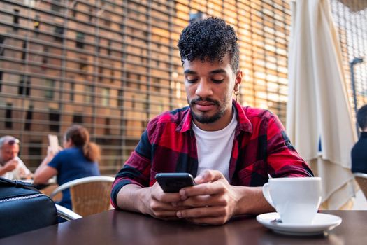 young latin man consulting his phone while sitting with a coffee on the terrace of a coffee shop, concept of technology and urban lifestyle