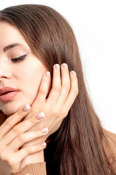 Close up portrait of pretty young woman touching her face by her hand with beige manicure on white background.