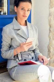 Woman is using smartphone while sitting near the door in the city, beautiful girl is holding a cell phone outdoor