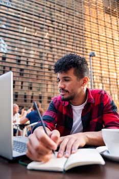 vertical photo of a young latin man studying with his computer and taking notes on the terrace of a coffee shop in the city, concept of technology and urban lifestyle