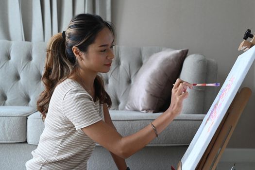 Relaxed young asian woman sitting in front of easel and painting picture with watercolor.