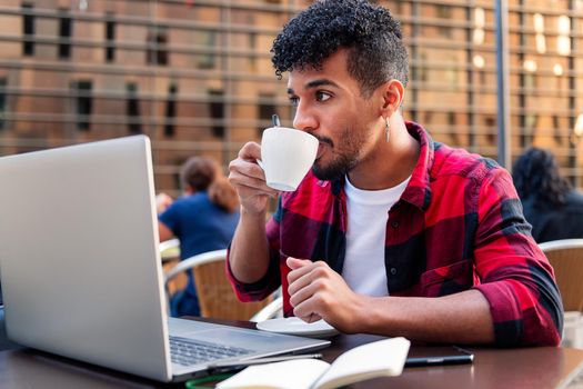 young latin man drinking coffee while working with his laptop on the terrace of a coffee shop, concept of technology and urban lifestyle