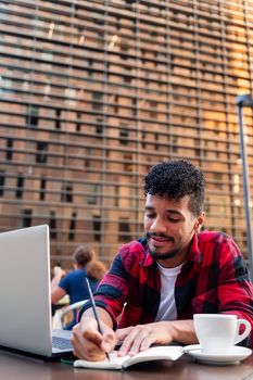 vertical photo of a young latin student studying with his computer and taking notes on the terrace of a coffee shop in the city, concept of technology and urban lifestyle