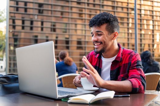 young latin man greets happy making a video call with his laptop from the terrace of a coffee shop, concept of technology and urban lifestyle