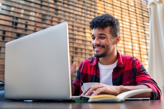 young latin man smiles making a video call with his laptop from the terrace of a coffee shop, concept of technology and urban lifestyle