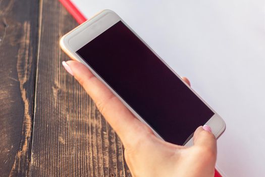 Mockup image of a woman's hands holding white mobile phone with blank black screen on background of white sheet of paper at a cafe.