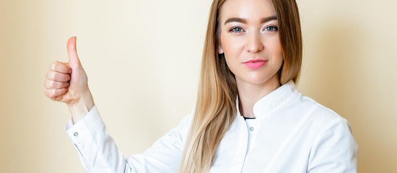 Beautiful young woman is showing thumbs up wearing white shirt over yellow background looking at camera.