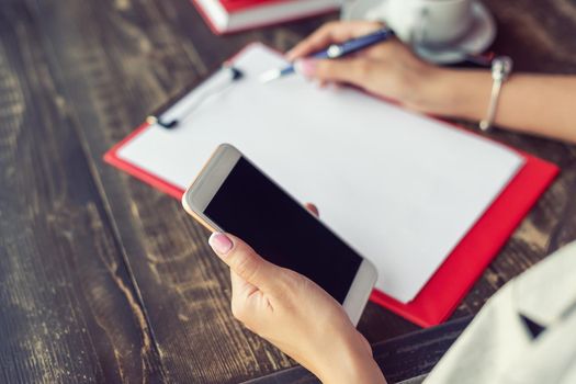 Mockup image of a woman's hands holding white mobile phone with blank black screen on background of white sheet of paper at a cafe.