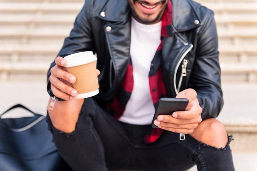 detail of the hands of an unrecognizable happy young man consulting the phone with a coffee in his hand, concept of technology and urban lifestyle