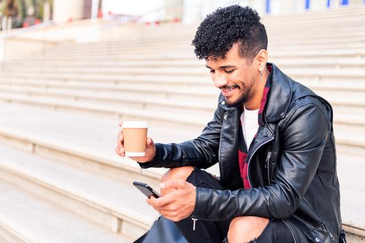 happy young latin student sitting on the stairs smiles consulting his phone with a coffee in his hand, concept of technology and urban lifestyle