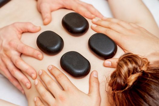 Male four hands laying large new oval black hot stones for back massage on female back in spa salon