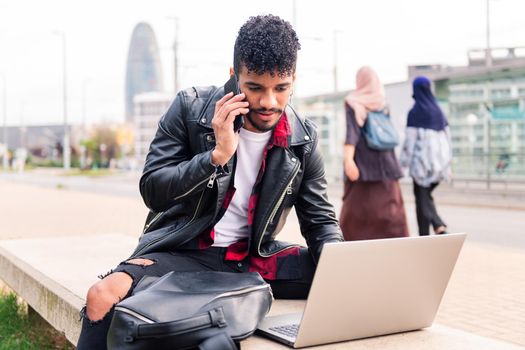 stylish latin man typing on his computer while talking on the phone sitting in the city as two arab women walk past him, concept of technology and urban lifestyle