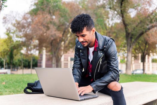 stylish latin student typing on his computer sitting on a park bench in the city, concept of technology and urban lifestyle